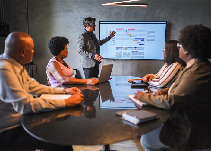 A professional is pointing at a computer screen showcasing a graph, offering a presentation to a group of individuals. They are seated across a table looking at the presentation.