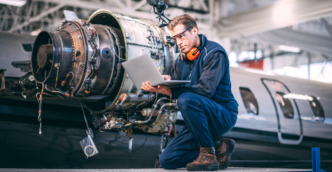 Aerospace engineer in coveralls and safety gear inspecting a jet engine with a laptop in a hangar