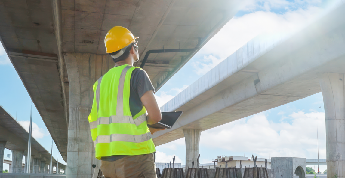 Construction engineer reviewing overhead cost allocation on a tablet at a bridge job site