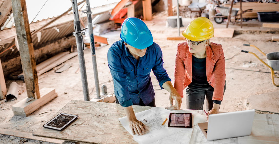 Architect and construction worker analyzing project progress using blueprints and a laptop on site