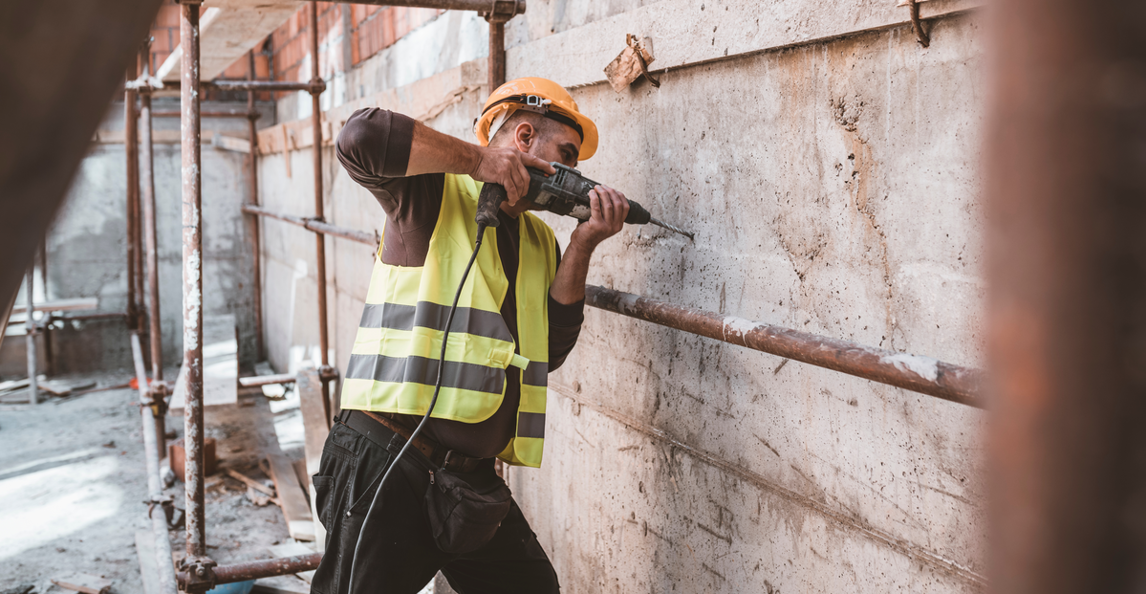 Construction worker in hard hat and hi-vis vest drilling into a concrete wall on a scaffold site