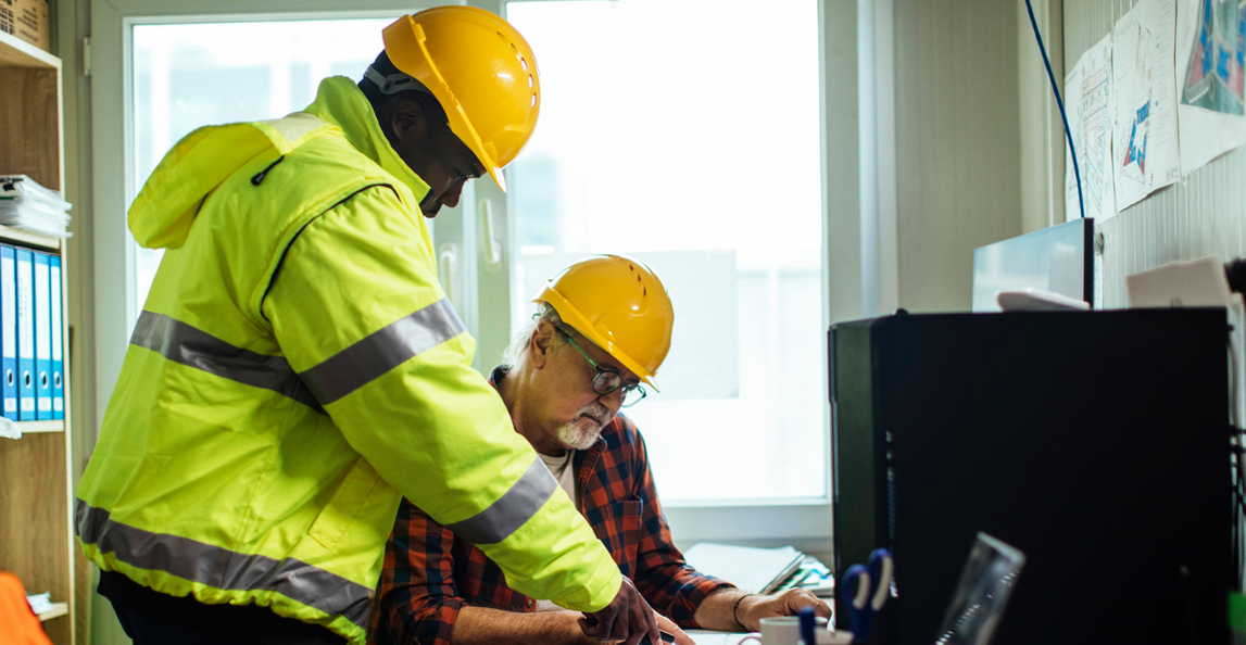 Two construction workers reviewing project payroll documents at a site office workstation