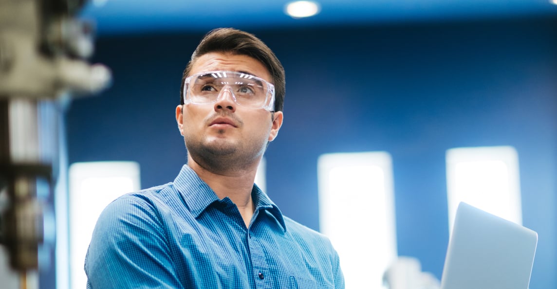 Manufacturing engineer in safety glasses using a laptop while inspecting machinery on a factory floor