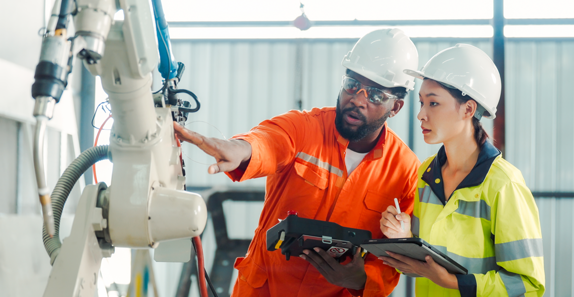 Two engineers in PPE inspecting a robotic arm and logging data on a tablet on a factory shop floor