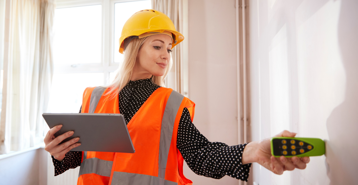 Quality inspector in orange safety vest and yellow hard hat operating a handheld device with a tablet in hand.