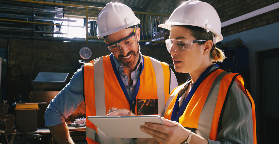 Two industrial workers in hard hats reviewing project data on a tablet in a manufacturing facility