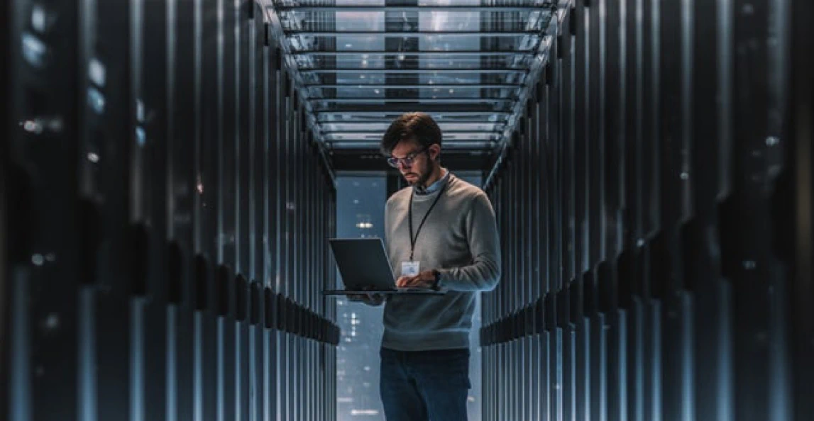 IT engineer working on a laptop in a data center aisle between rows of server racks