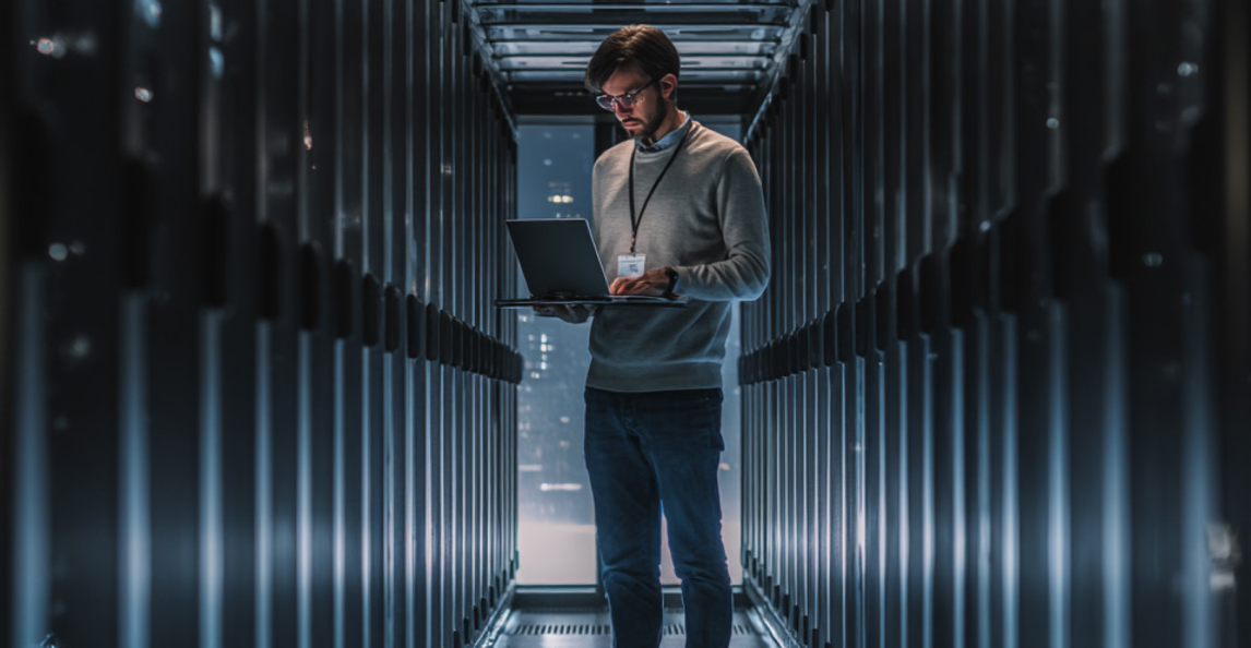 An IT professional with a laptop stands in a secure data centre corridor.