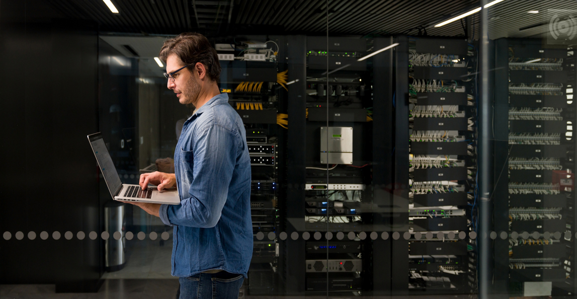 An IT technician works on a laptop in a glass-walled server room.