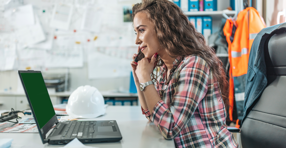 Construction estimator using a laptop in a site office with blueprints and hard hat
