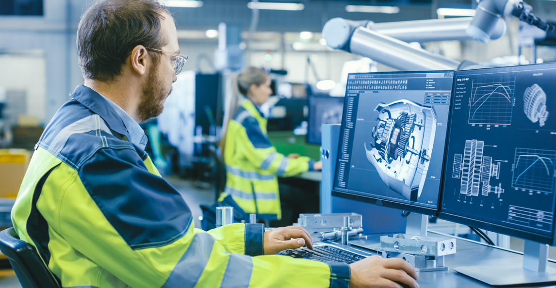 Manufacturing engineer in hi-vis vest reviewing a 3D CAD model on dual monitors in a factory