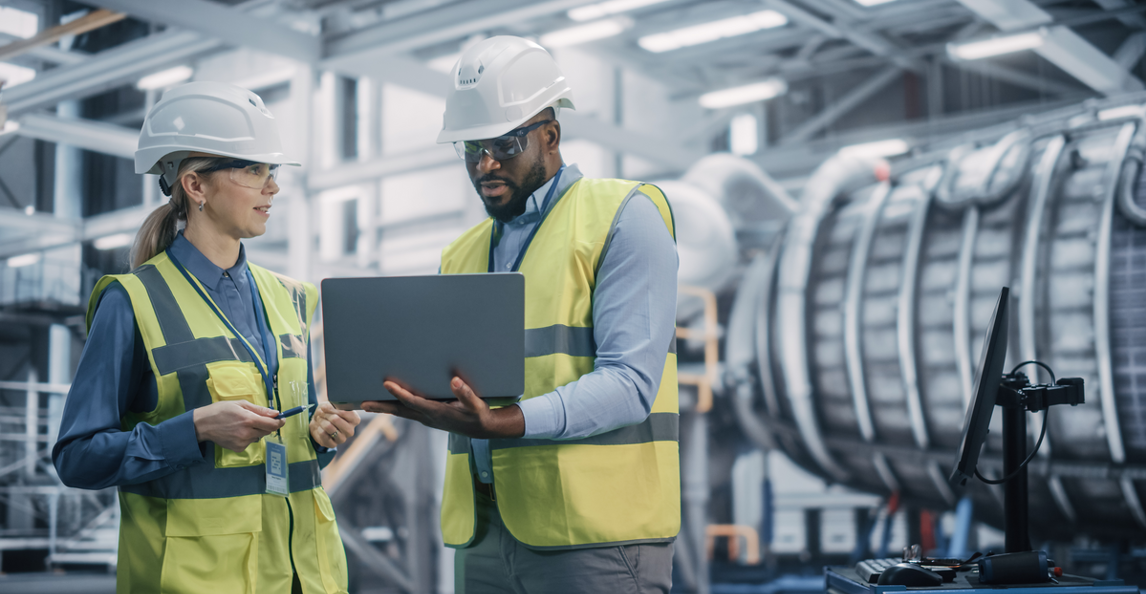 Two engineers in hi-vis vests reviewing a laptop on a factory floor