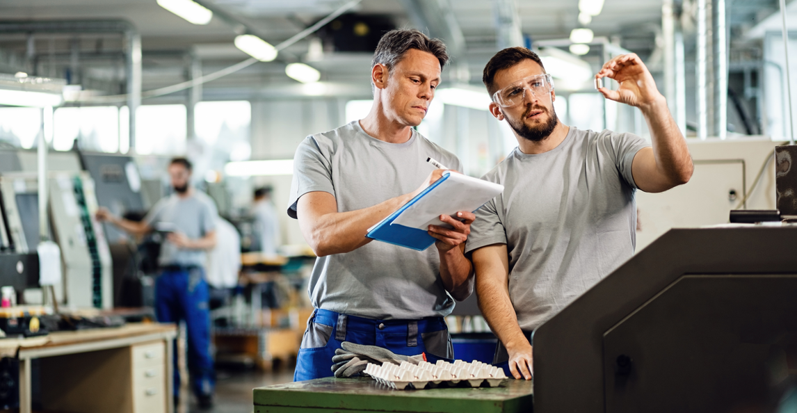 Two workers inspect a machined component on a factory floor.