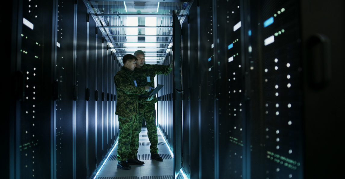 Two military personnel reviewing data on a laptop in a data center