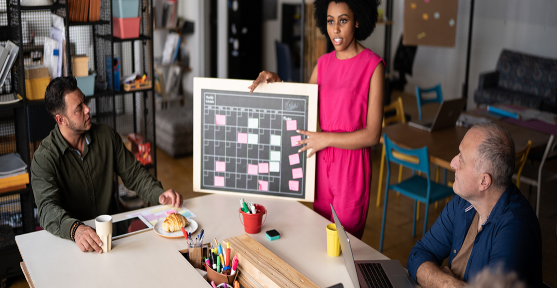 Project manager presenting a scheduling board with sticky notes to a team in a meeting room