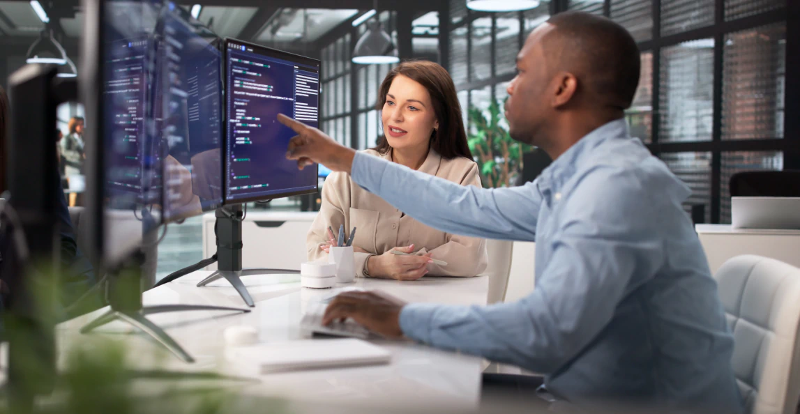 Two professionals collaborate at a workstation, reviewing software code on dual monitors in a modern office environment
