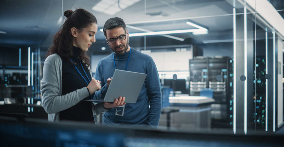 Two IT professionals review a laptop in a modern glass-walled data centre.