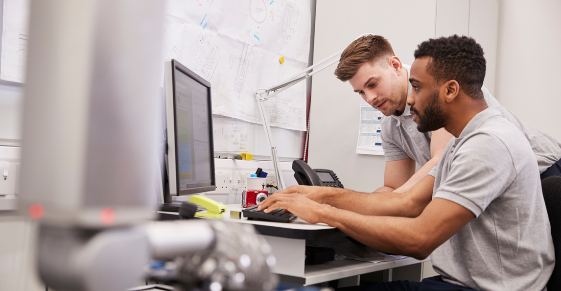 Two professionals collaborating at a computer workstation with technical plans displayed in the background.