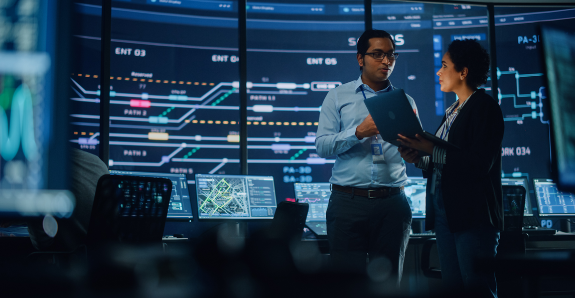 Two professionals discussing project data in a high-tech government system monitoring control room.
