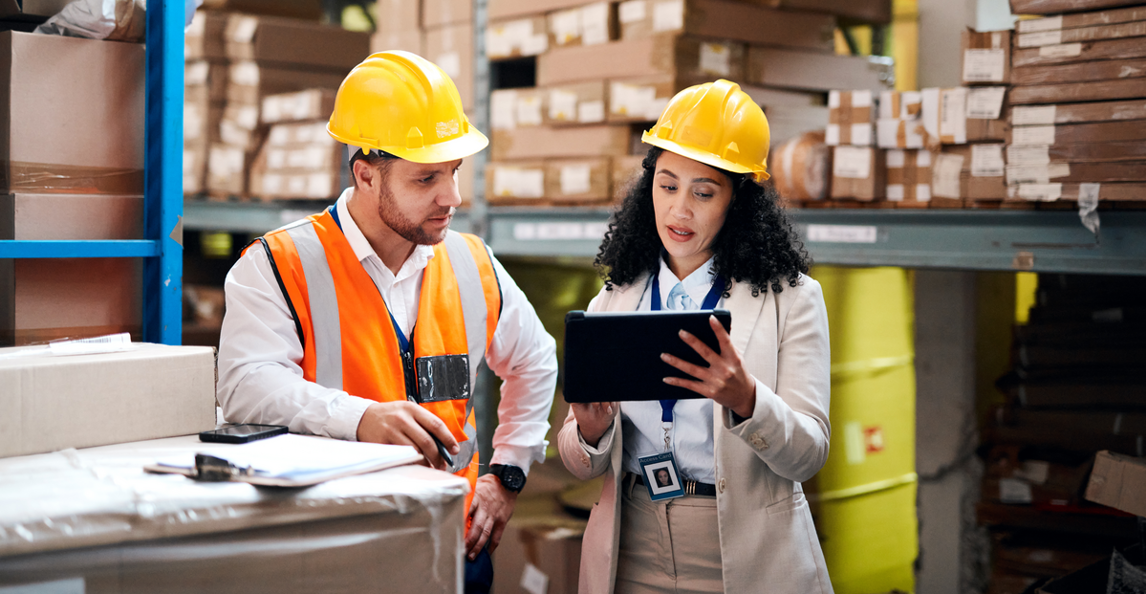 Two warehouse workers in hard hats reviewing incoming goods data on a tablet