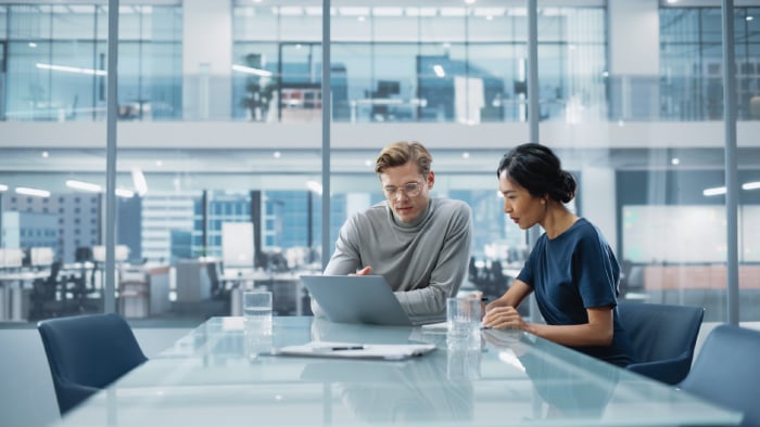 Two Business Professionals Reviewing Project Data and Reports on Laptop in Modern Corporate Office