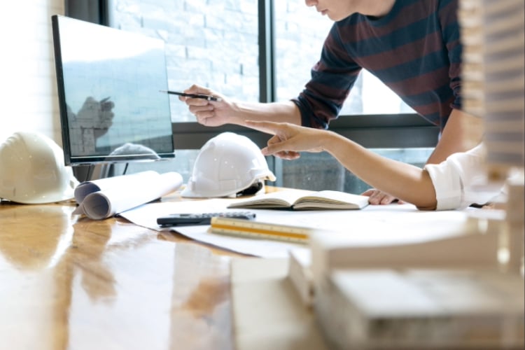 Two contractors reviewing project plans and documents at a desk with hard hats, blueprints, and a computer monitor