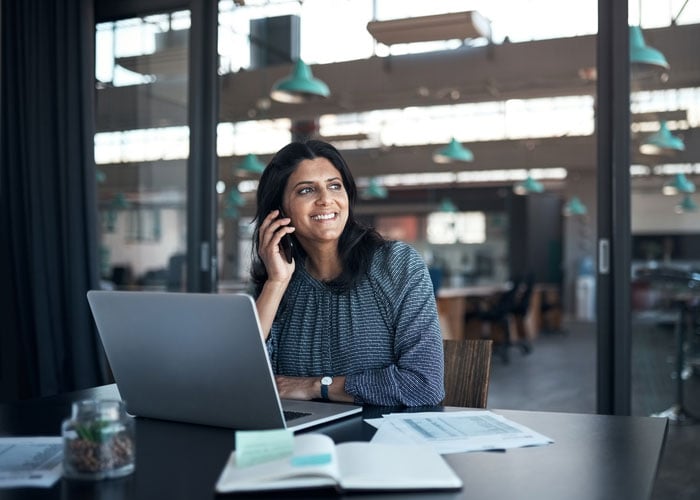 Business Professional Managing Client Communication on Phone while Working on Laptop in Modern Office