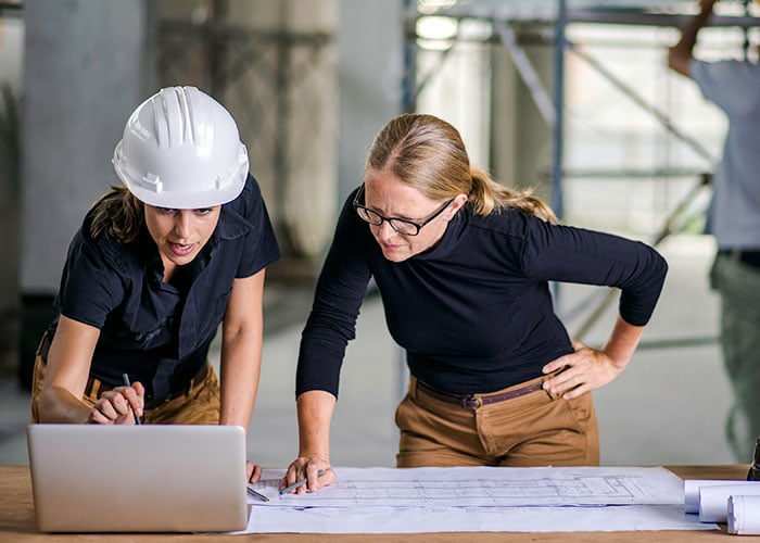 Front View of two women standing by the desk full of blueprint and laptop construction worker in the background
