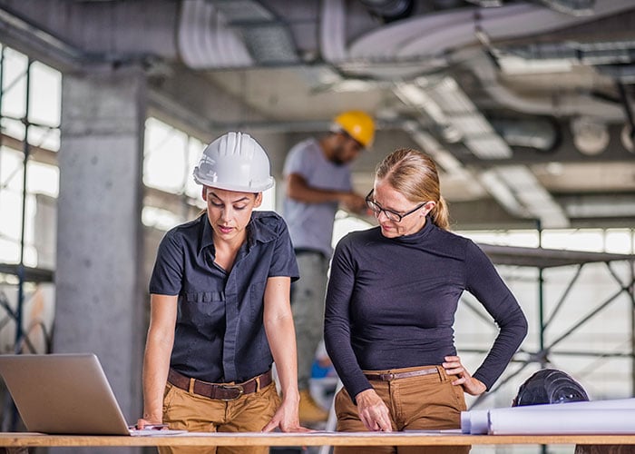 Two women standing by a desk full of blueprints and a laptop